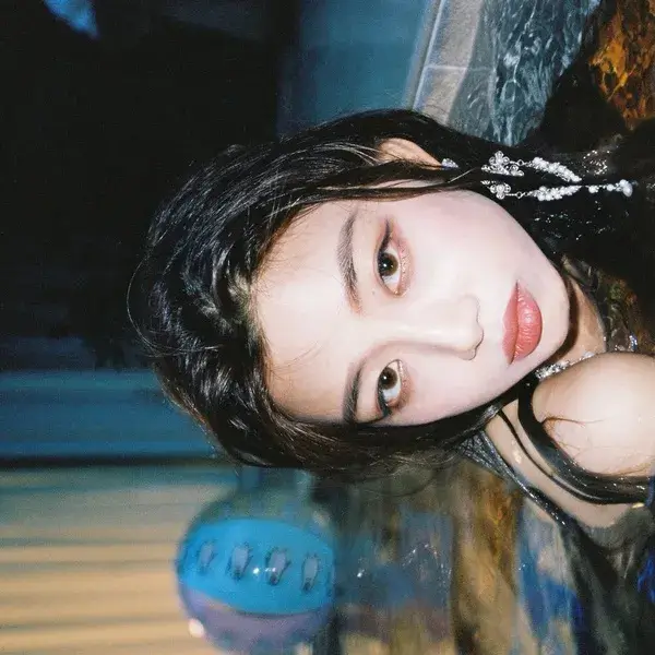 한 인물이 물속에 반쯤 잠긴 채 옆으로 누워 카메라를 응시하는 콘셉트 사진. 화면이 90도 회전된 구도로, 젖은 검은 머리가 수면 위로 퍼져 있고 이마와 목에는 은빛 장식이 물기와 함께 반짝인다. 푸른 조명이 물 전체를 감싸며 아래쪽에는 흔들리는 반영이 번져 있다. 현실과 물속 사이 경계에 놓인 듯한 몽환적인 분위기가 인상적이다. 숀 Way Back Home
